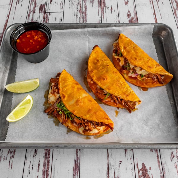 Three Calle Taco birria tacos with lime wedges and a side of consommé on a metal tray.